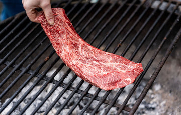 A raw, whole grass fed beef flat iron steak being laid onto a barbecue