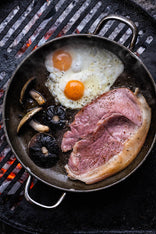 A native breed unsmoked gammon pork steaks cooking in a pan with mushrooms and eggs on a barbecue.