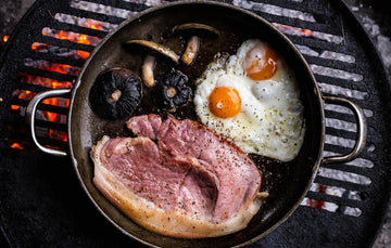 A native breed unsmoked gammon pork steak cooking in a pan with mushrooms and eggs on a barbecue.
