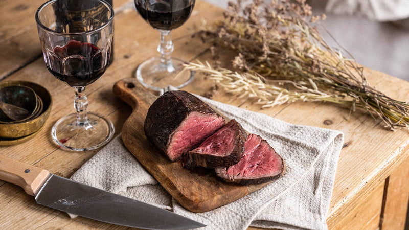 A cooked and sliced grass fed Beef Chateaubriand sharing steak with glasses of wine and dried flowers.