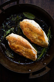 Two properly free range chicken breasts cooking in a pan with fresh herbs.